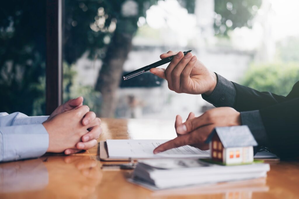A female attorney holding a pen and pointing at paperwork across from a client at a desk next to a model house.